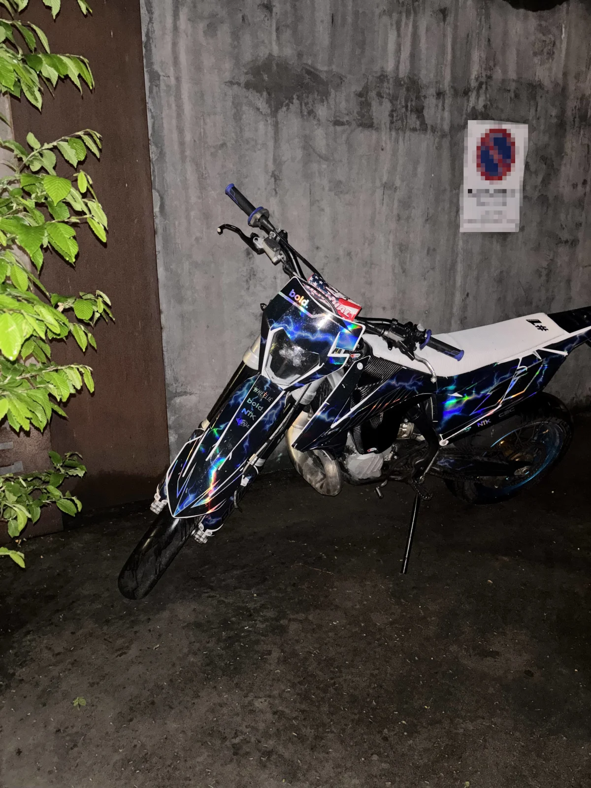 Ein Motorrad mit buntem Muster ist an einer Betonwand abgestellt. Ein Schild an der Wand ist unscharf erkennbar.