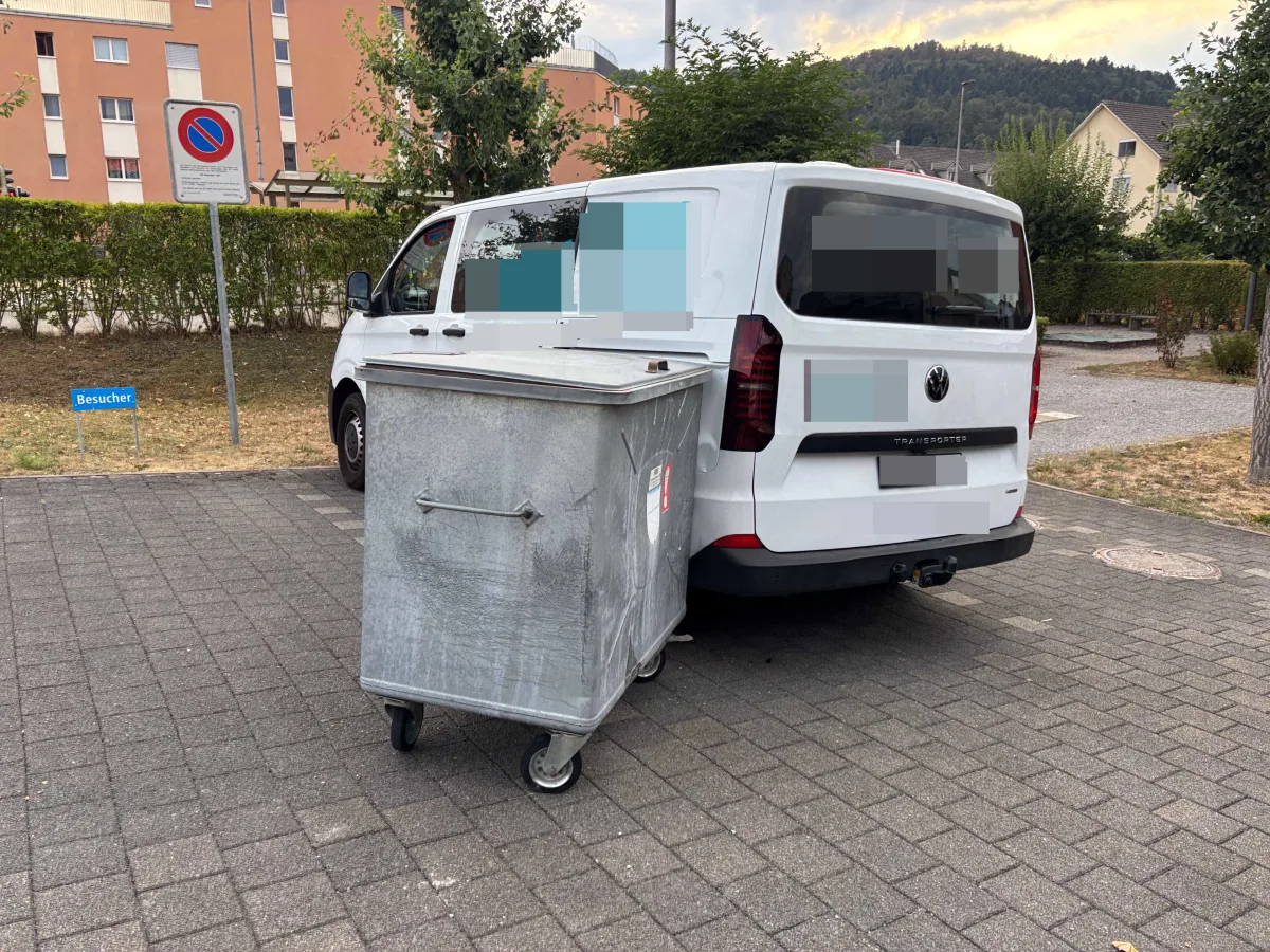Auf dem Bild ist ein weißer VW-Transporter zu sehen, der auf einem Parkplatz neben einem großen Müllcontainer steht. Der Hintergrund zeigt Wohngebäude und Bäume.
