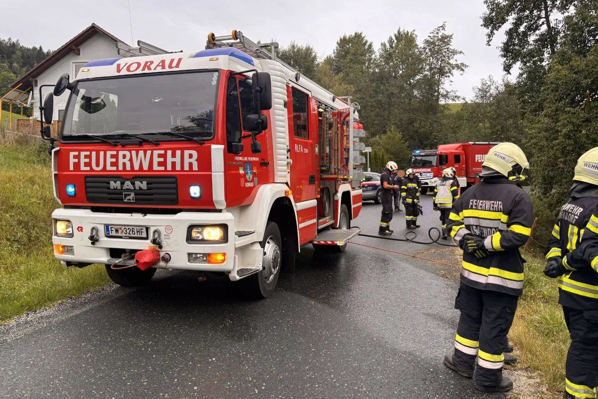 Ein rotes Feuerwehrauto der Feuerwehr Vorau steht auf einer Landstraße. Einsatzkräfte in Uniformen sind im Hintergrund zu sehen.