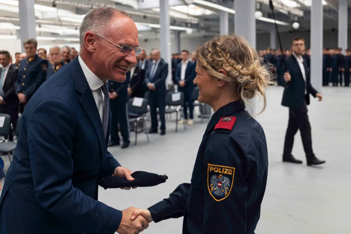 In einer großen Halle schütteln sich eine Polizistin in Uniform und ein Mann im Anzug die Hand. Im Hintergrund sind mehrere Personen in Uniformen zu sehen.