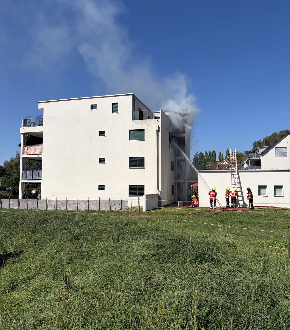 Ein mehrstöckiges Wohngebäude mit Rauch, der aus dem oberen Stockwerk aufsteigt. Feuerwehrleute sind mit einem Löschschlauch im Einsatz.