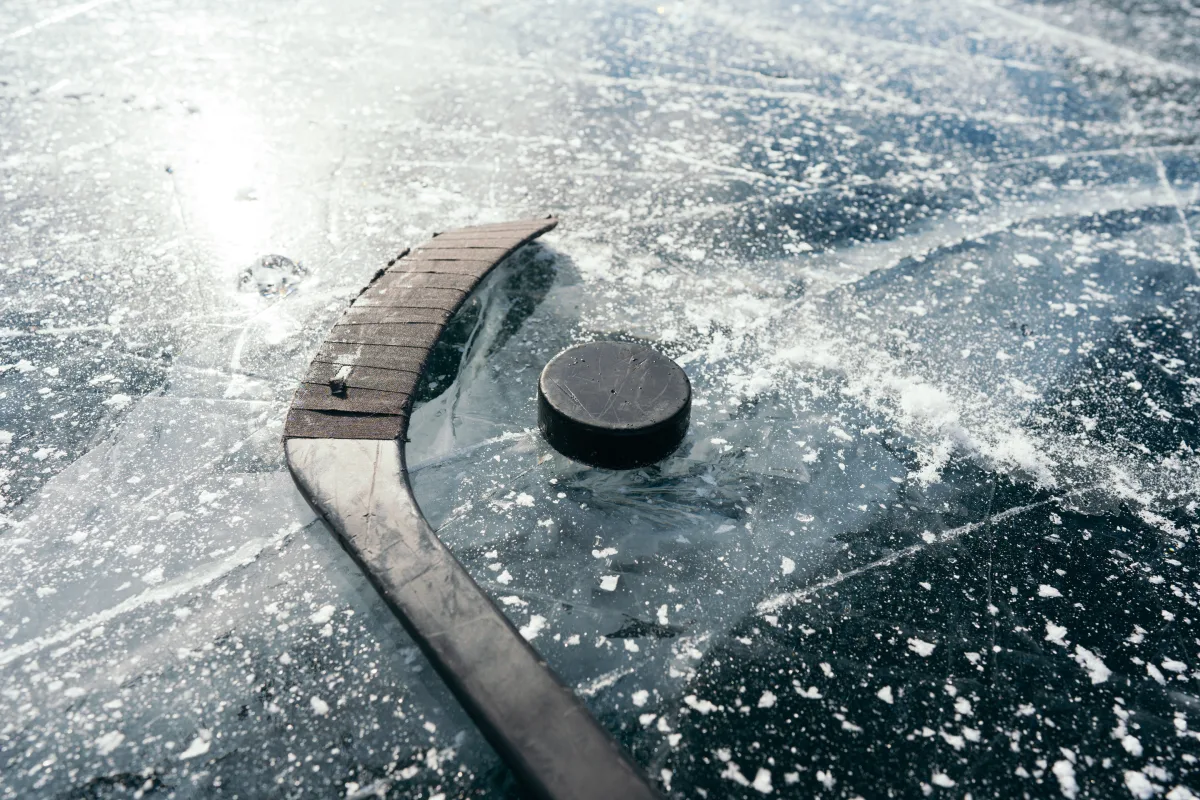 Ein Hockeyschläger und ein Puck liegen auf einer glatten, vereisten Fläche mit sichtbaren Kratzspuren und leichtem Schneebelag.