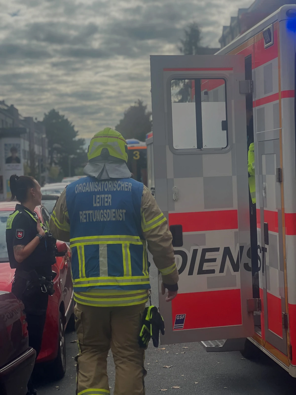 Ein Mitarbeiter des Rettungsdienstes steht neben einem geöffneten Einsatzfahrzeug, während eine weitere Person in Uniform danebensteht. Es ist bewölkt und ein weiteres Fahrzeug ist im Hintergrund sichtbar.
