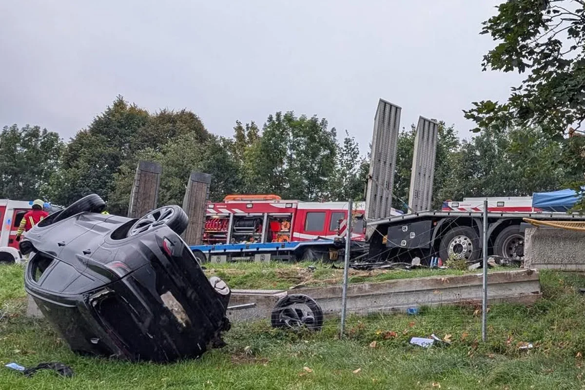 Ein Auto liegt umgestürzt neben Feuerwehrfahrzeugen auf einer grasbewachsenen Fläche, umgeben von Bäumen.