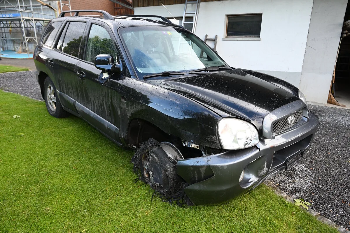 Ein schwarzes Auto mit einem stark beschädigten und entblößten Vorderreifen steht auf einem Rasenstück. Es hat sichtbare Schäden an der Karosserie.