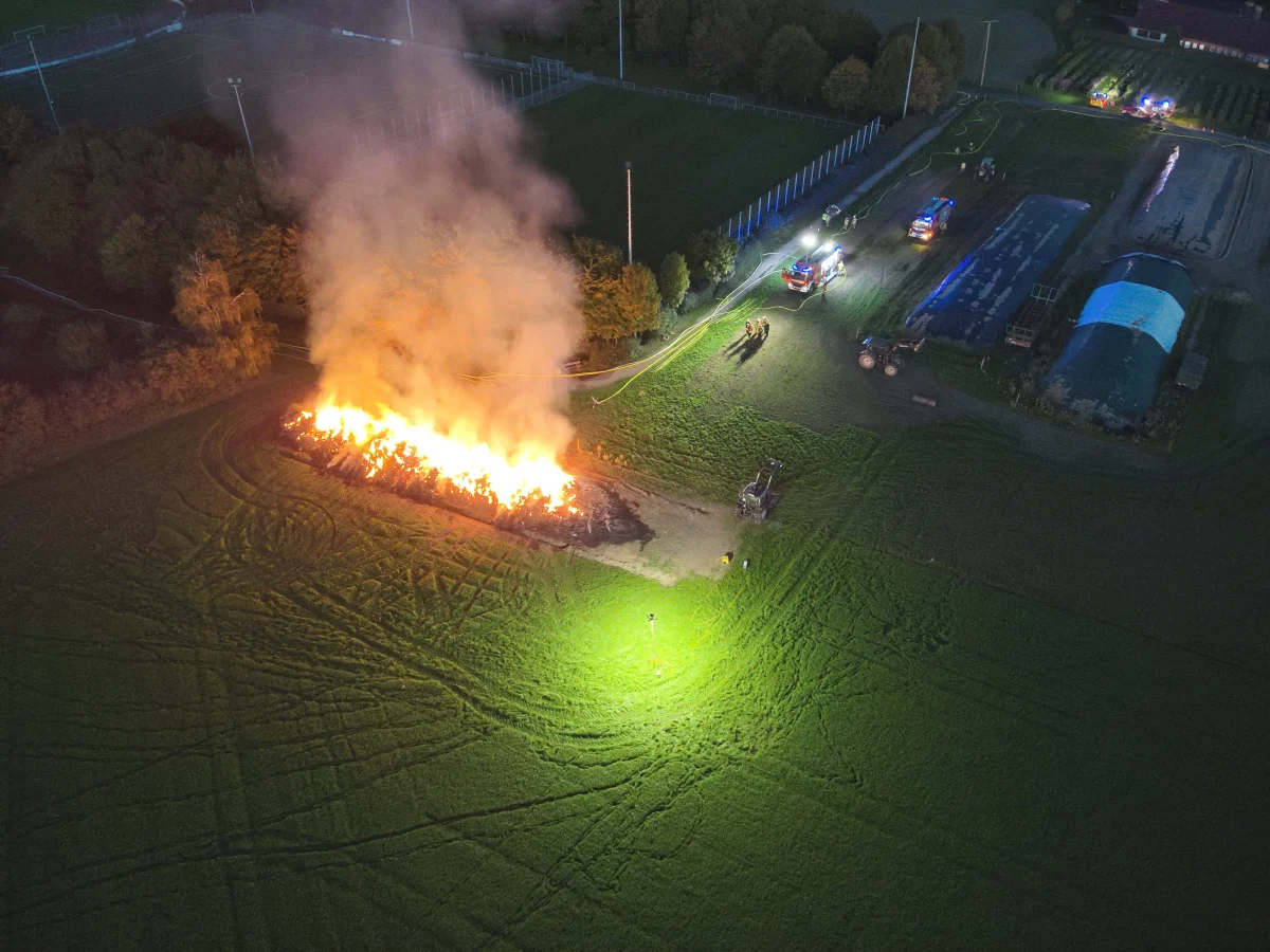 Eine große Feuerstelle brennt auf einem Feld bei Nacht. Rundherum sind Einsatzfahrzeuge mit blinkenden Lichtern sichtbar.
