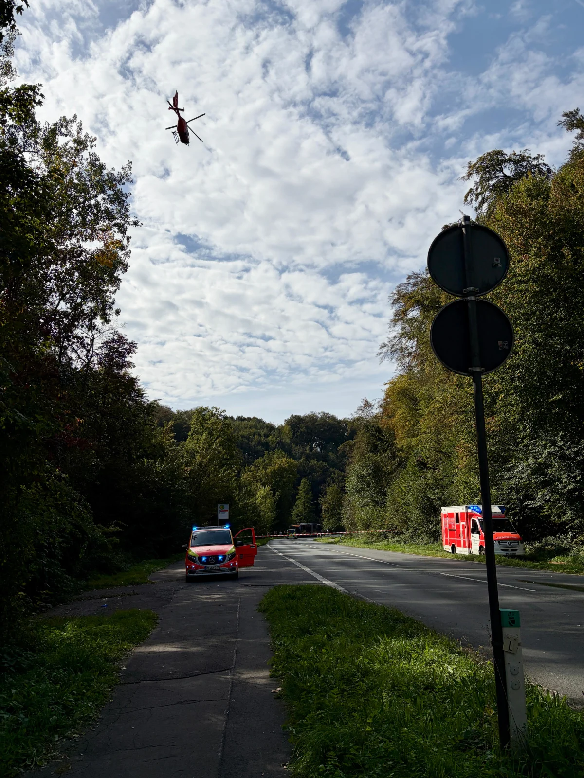 Ein roter Rettungshubschrauber fliegt über einer von Wald gesäumten Landstraße. Polizei- und Rettungsfahrzeuge sind auf der Straße zu sehen.