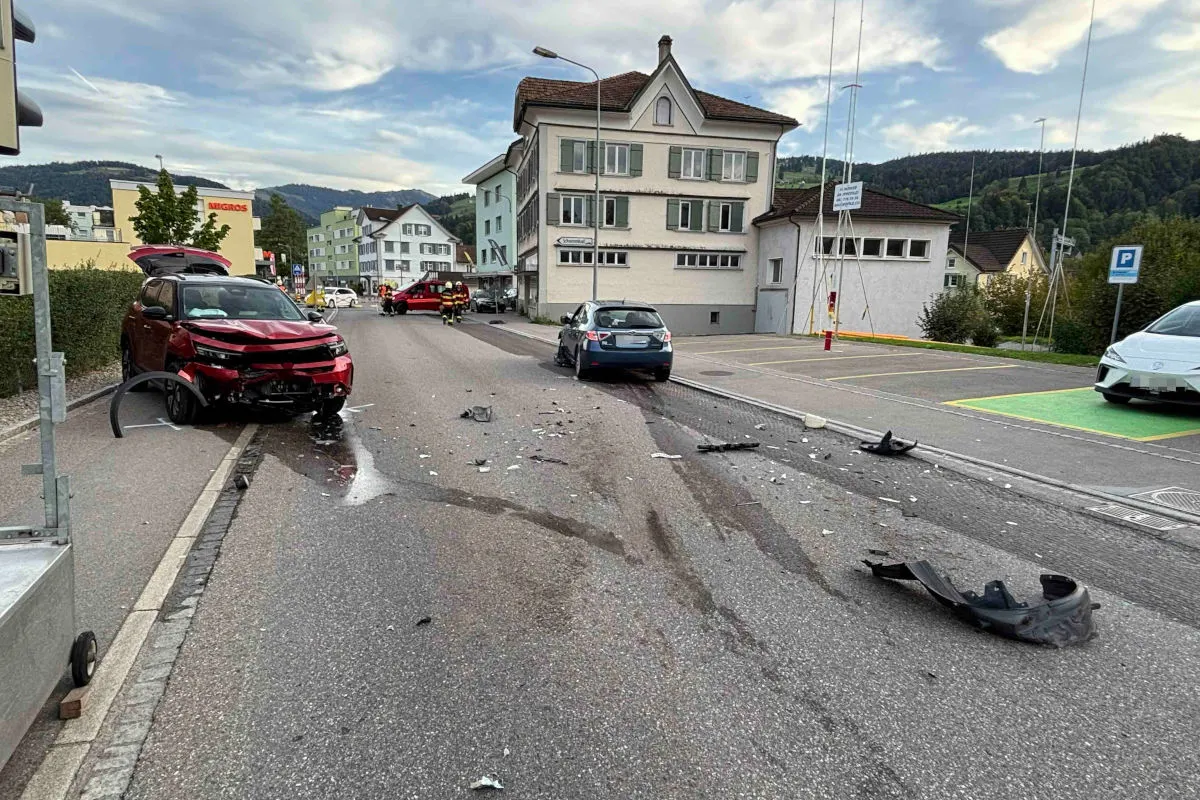 Ein rotes Auto mit erheblichen Frontschäden steht auf einer Straße, umgeben von Trümmerteilen. Im Hintergrund sind Wohnhäuser und Berge sichtbar.