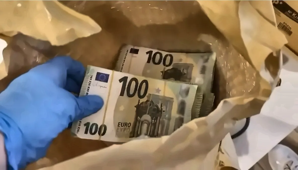 Eine Person mit blauen Handschuhen hält mehrere 100-Euro-Scheine in einer braunen Papiertüte. Die Szene findet scheinbar in einem Innenraum statt.