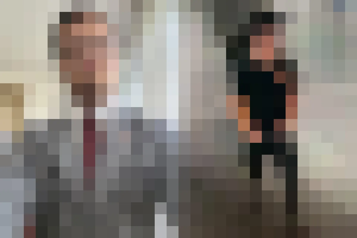 Eine Person trägt einen Anzug und steht vor einem neutralen Hintergrund. Die zweite Person trägt Freizeitkleidung in einem Innenraum.