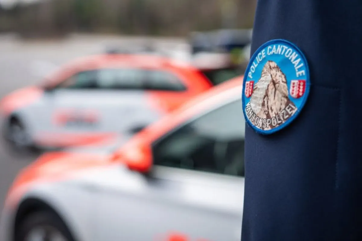 Im Vordergrund ist ein Abzeichen der Kantonspolizei Wallis auf einer Uniform zu sehen; im Hintergrund unscharfe Polizei-Einsatzfahrzeuge.