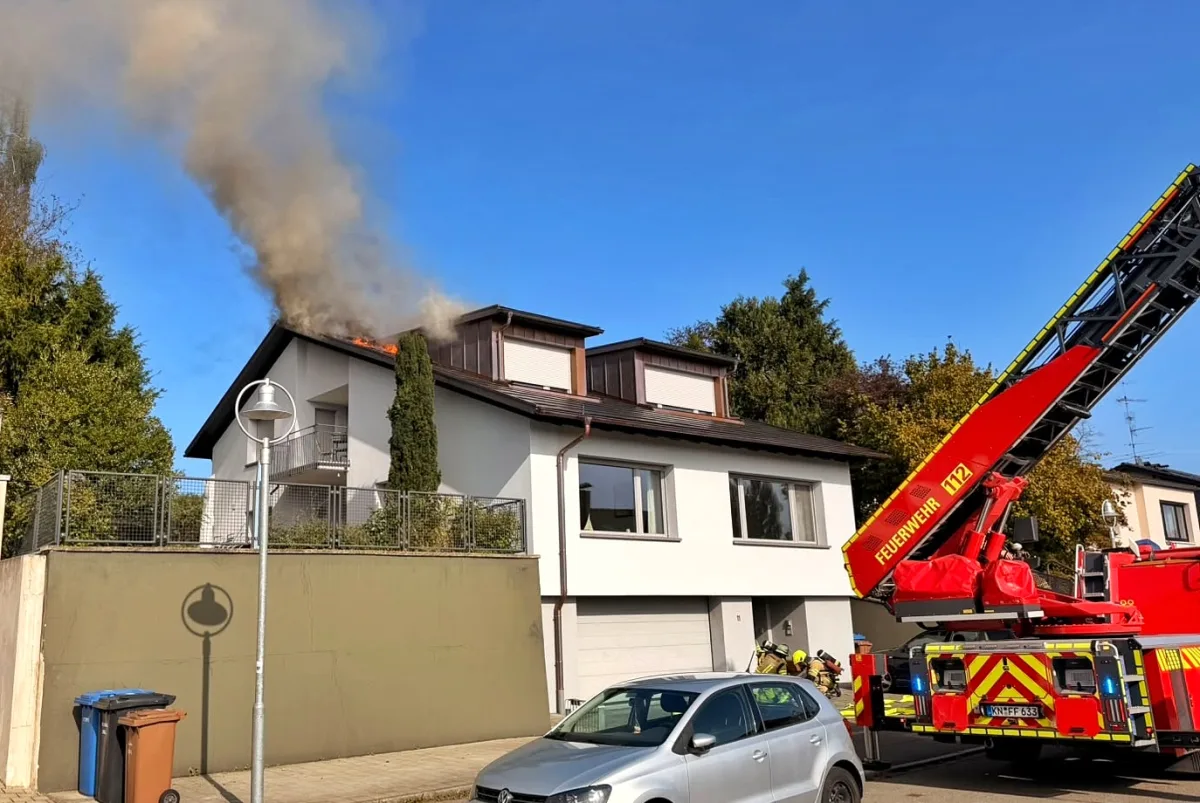 Ein Hausdach brennt und Rauch steigt empor. Eine Feuerwehrleiter ist ausgefahren, und mehrere Einsatzkräfte sind anwesend.