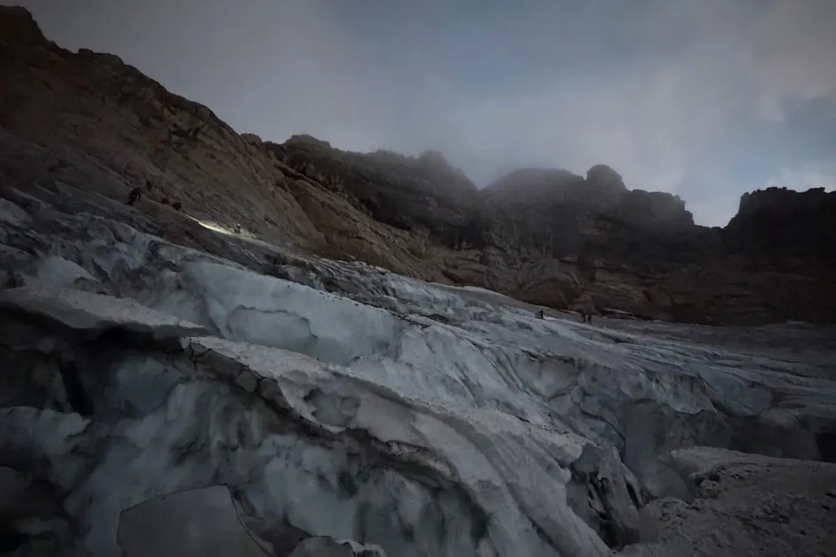 Ein Gletscher im Hochgebirge bei Dämmerung, auf dem Bergsteiger mit Taschenlampen unterwegs sind. Die dunkle Szenerie betont die Dramatik der Suche.