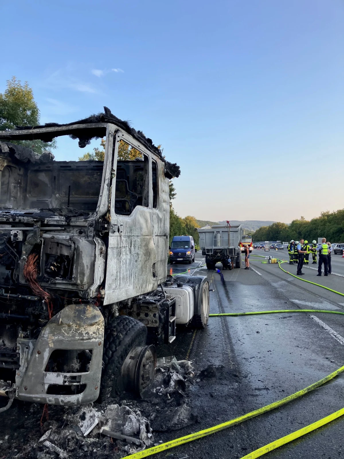 Ein ausgebrannter Lastwagen steht auf der Autobahn, umgeben von Polizeikräften. Verkehrssicherungsschläuche sind ausgelegt.