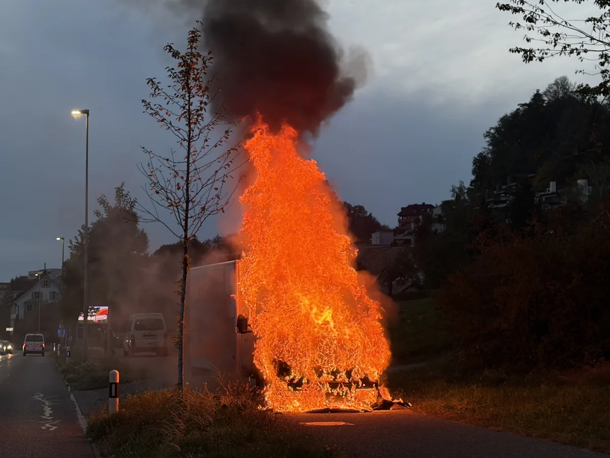 Ein Fahrzeug steht am Straßenrand in Flammen, begleitet von intensiver Rauchentwicklung. Die Szene befindet sich in einer städtischen Umgebung bei Dämmerung.