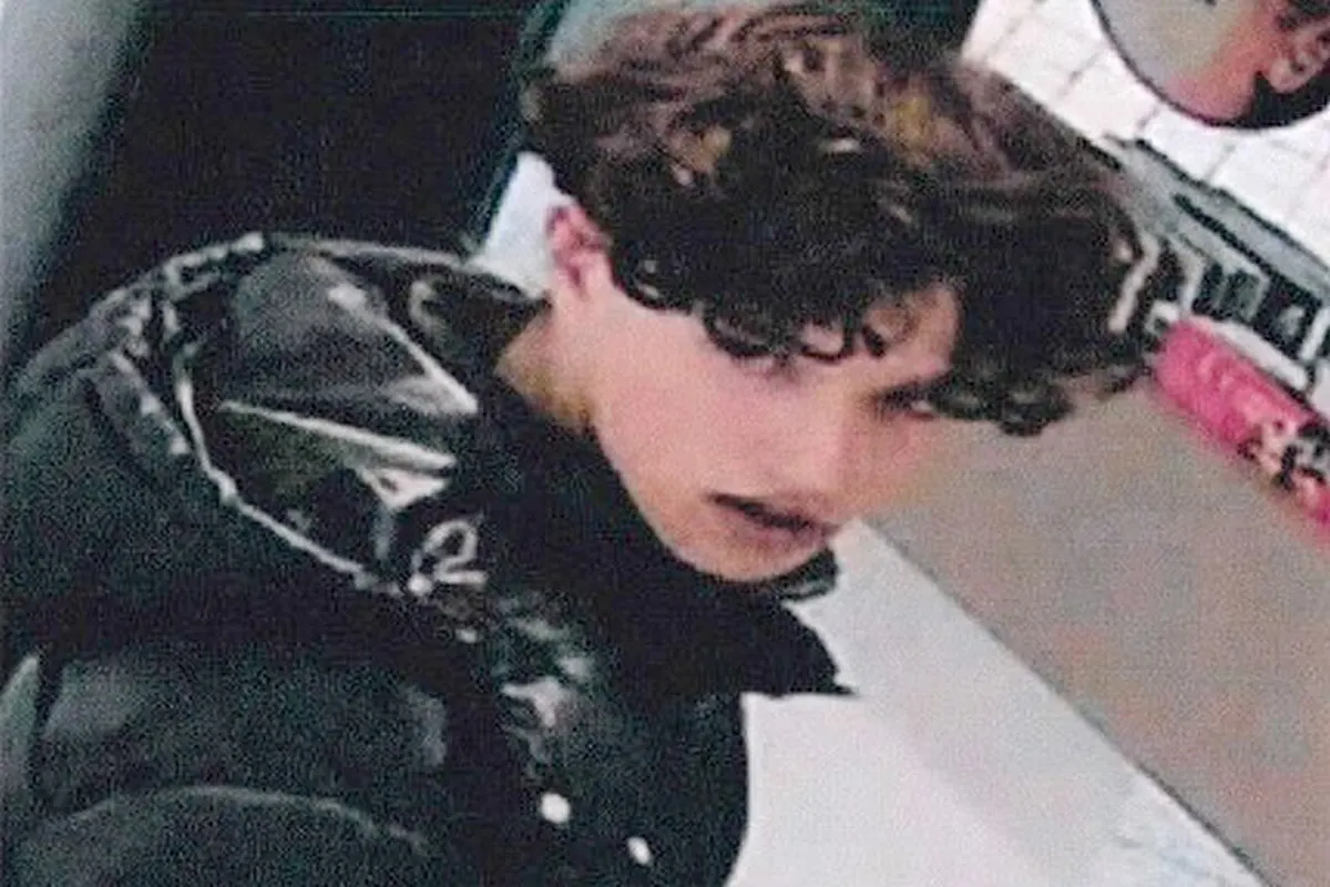 Eine junge Person mit lockigem Haar und dunkler Kleidung wird von einer Überwachungskamera in einem Innenraum aufgenommen. Im Hintergrund sind Regale zu sehen.