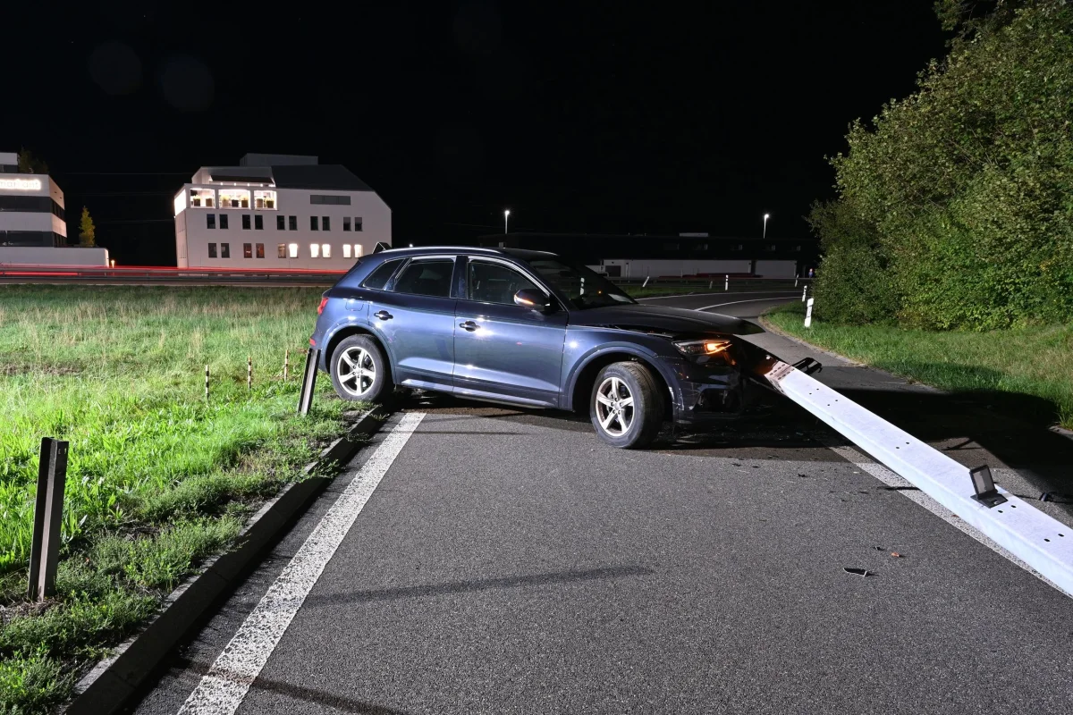 Ein blauer SUV steht mit der Front in einer Leitplanke auf einer Landstraße. Die Szene ist nachts, mit beleuchteten Gebäuden im Hintergrund.