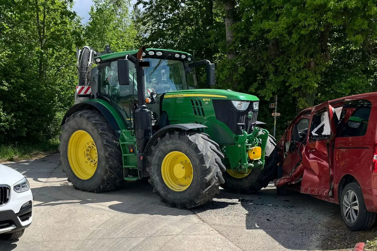 Ein grüner Traktor hat ein mit einem roten Auto auf einer Landstraße kollidiert. Beide Fahrzeuge weisen sichtbare Schäden auf, und die Umgebung ist ländlich mit vielen Bäumen.