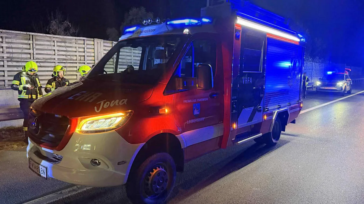 Ein rotes Feuerwehrfahrzeug, beschriftet mit 'Freiwillige Feuerwehr', steht mit eingeschaltetem Blaulicht auf einer Straße. Drei Feuerwehrleute in Einsatzkleidung sind daneben zu sehen.