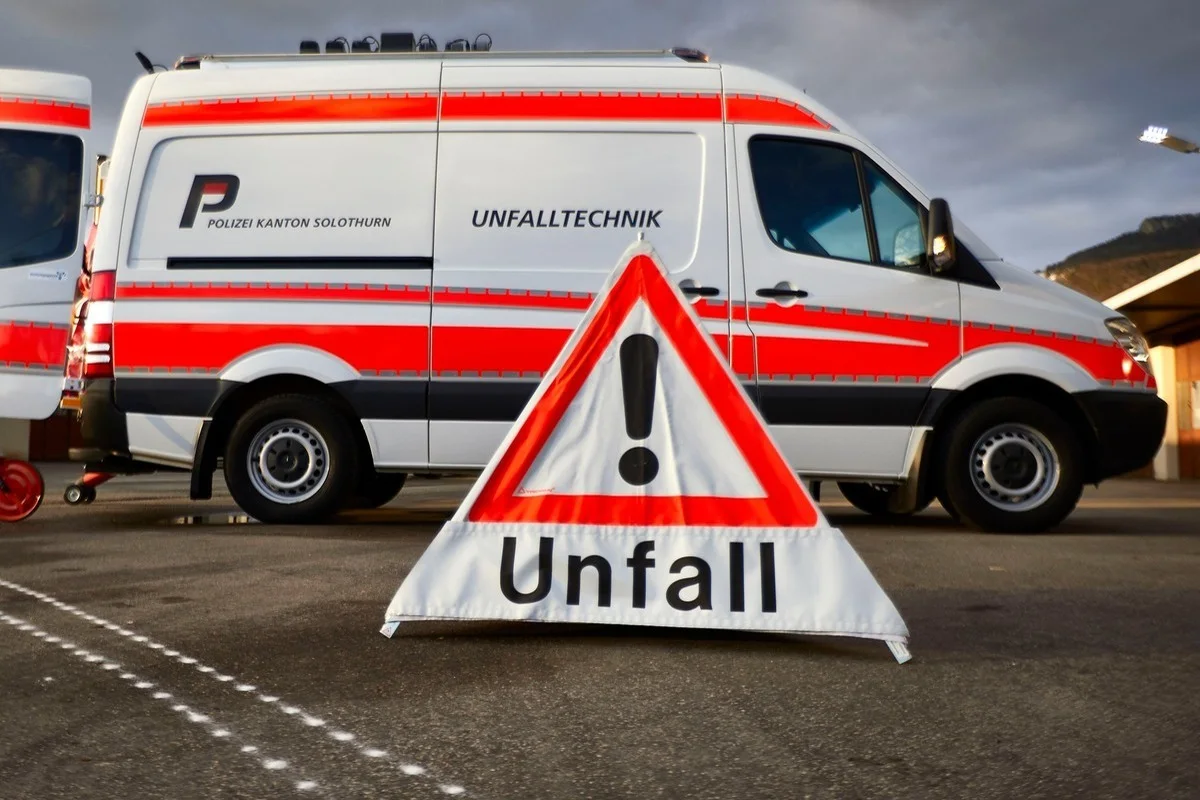 Im Vordergrund steht ein Warndreieck mit einem Ausrufezeichen und dem Wort 'Unfall'. Dahinter ist ein Fahrzeug der Kantonspolizei Solothurn mit der Aufschrift 'Unfalltechnik' abgebildet.