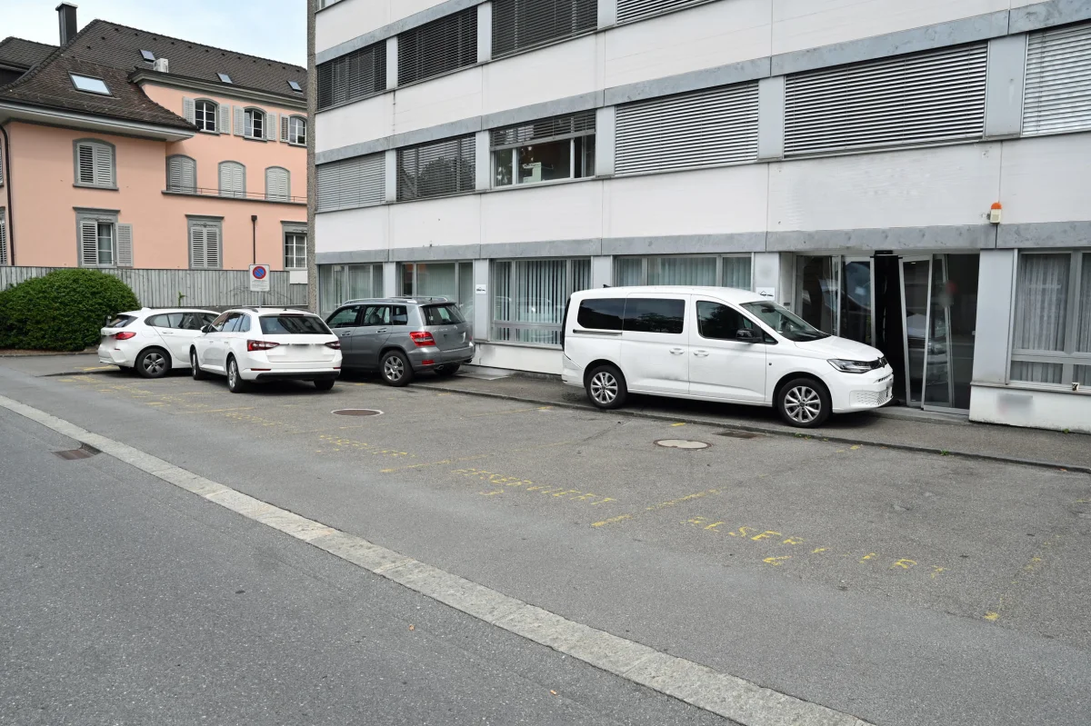 Ein weißer Van und zwei Kombis stehen auf markierten Parkplätzen vor einem mehrstöckigen Bürokomplex. Im Hintergrund ist ein Wohngebäude sichtbar.