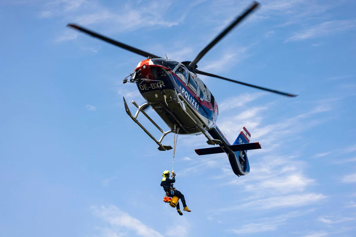 Ein Polizeihubschrauber schwebt am Himmel, ein Beamter seilt sich daran herab. Der Hintergrund ist ein klarer blauer Himmel.