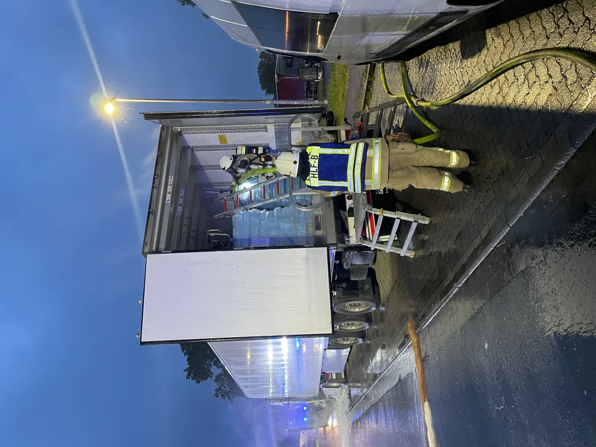 Ein Feuerwehrmann, gekleidet in Schutzkleidung, ist an einem LKW am Straßenrand bei Dunkelheit tätig. Der Himmel ist leicht bewölkt und ein Schlauch ist ausgerollt.