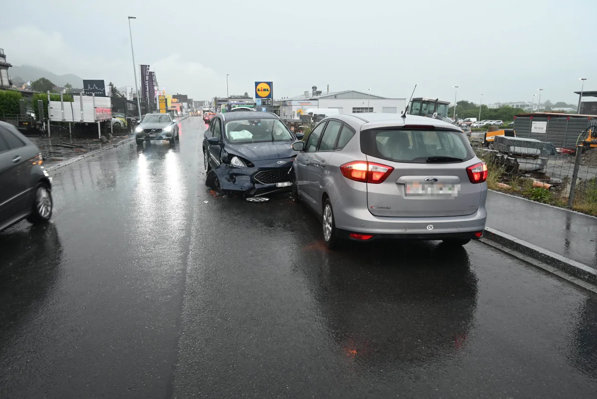 Zwei Fahrzeuge, ein blauer und ein grauer Ford, stehen nach einer Kollision auf einer regennassen Straße. Im Hintergrund ist ein Supermarkt zu sehen.