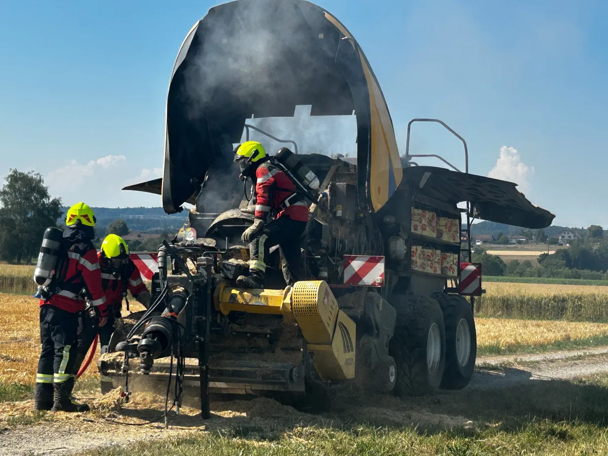 Mehrere Feuerwehrleute in Schutzkleidung löschen ein brennendes landwirtschaftliches Fahrzeug auf einem Feld bei klarem Wetter. Rauch steigt aus dem Fahrzeug auf.