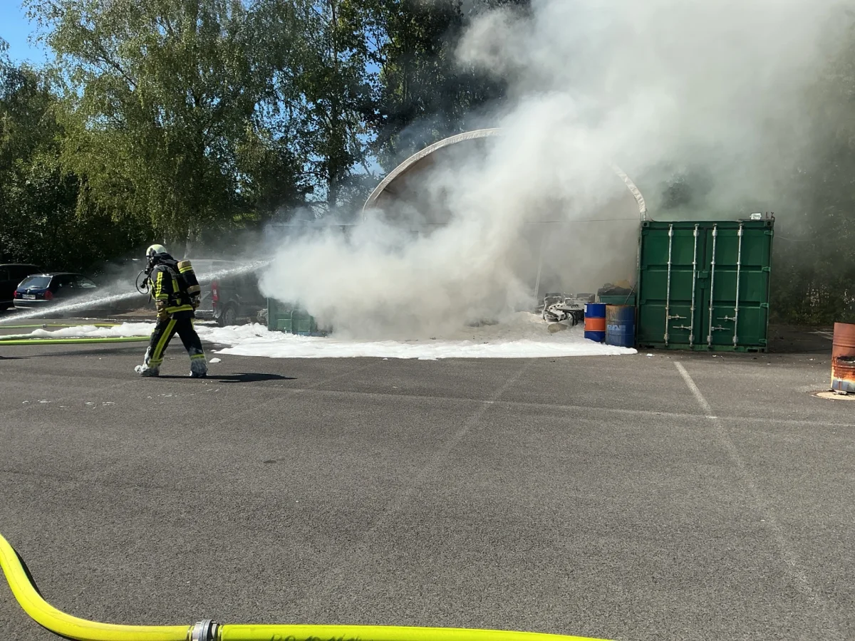 Ein Feuerwehrmann bekämpft einen Brand, indem er mit einem Wasserstrahl auf Rauch vor einem Container zielt. Dicker Rauch umhüllt die Umgebung.