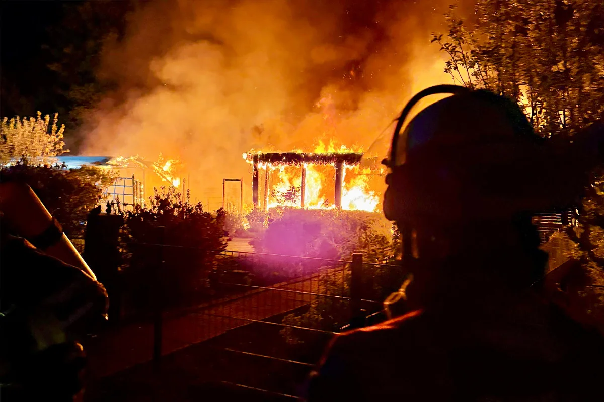 Ein Gebäude steht in Flammen bei Nacht. Im Vordergrund sind die Silhouetten von Feuerwehrleuten zu erkennen, die die Szene beobachten.