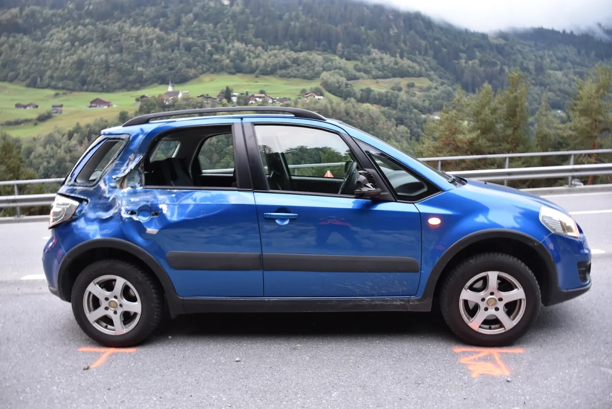 Ein blauer Pkw mit deutlichen Schäden an der Seite steht still auf einer Landstraße, umgeben von grüner Landschaft.
