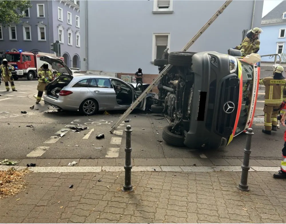 Ein umgekippter Krankenwagen liegt neben einem silbernen Kombi auf einer städtischen Straße. Feuerwehrleute sind vor Ort.