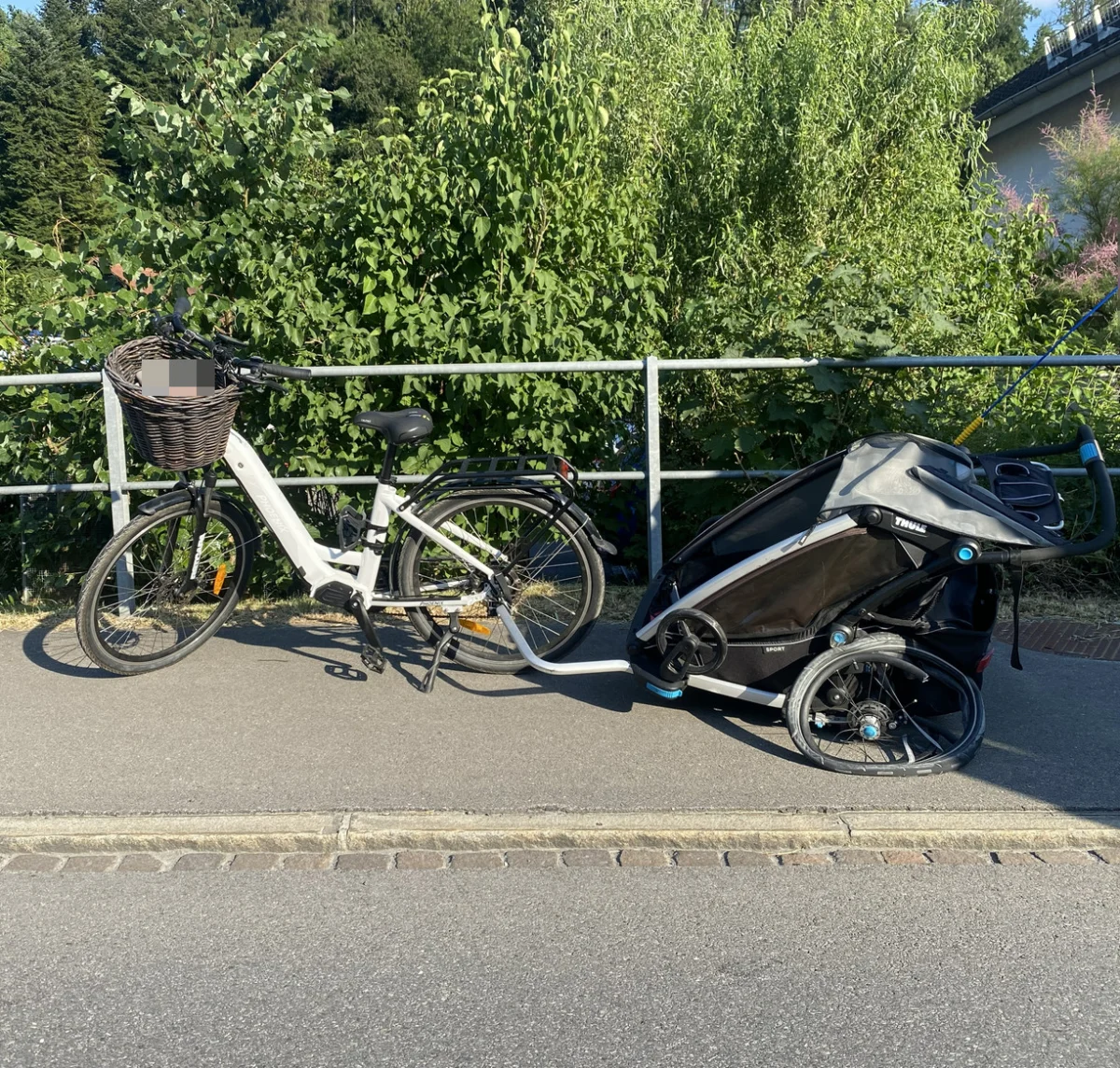 Ein weißes Fahrrad mit einem Korb vorne, angebunden an einen schwarzen Kinderanhänger, steht auf einem Gehweg. Im Hintergrund sind dicht bewachsene grüne Büsche sichtbar.