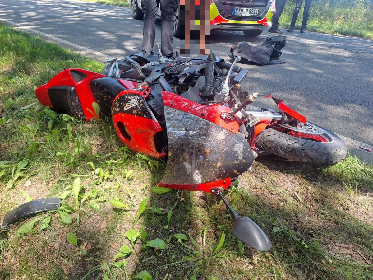 Ein rotes Motorrad liegt beschädigt im Gras neben einer Straße, während im Hintergrund ein Polizeifahrzeug sichtbar ist.