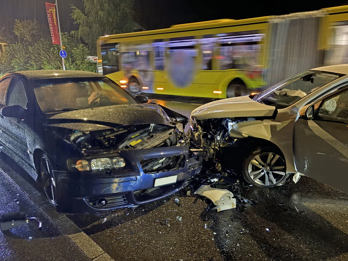 Ein schwarzes und ein silbernes Auto kollidierten frontal auf einer nassen Straße. Im Hintergrund fährt ein gelber Bus vorbei.
