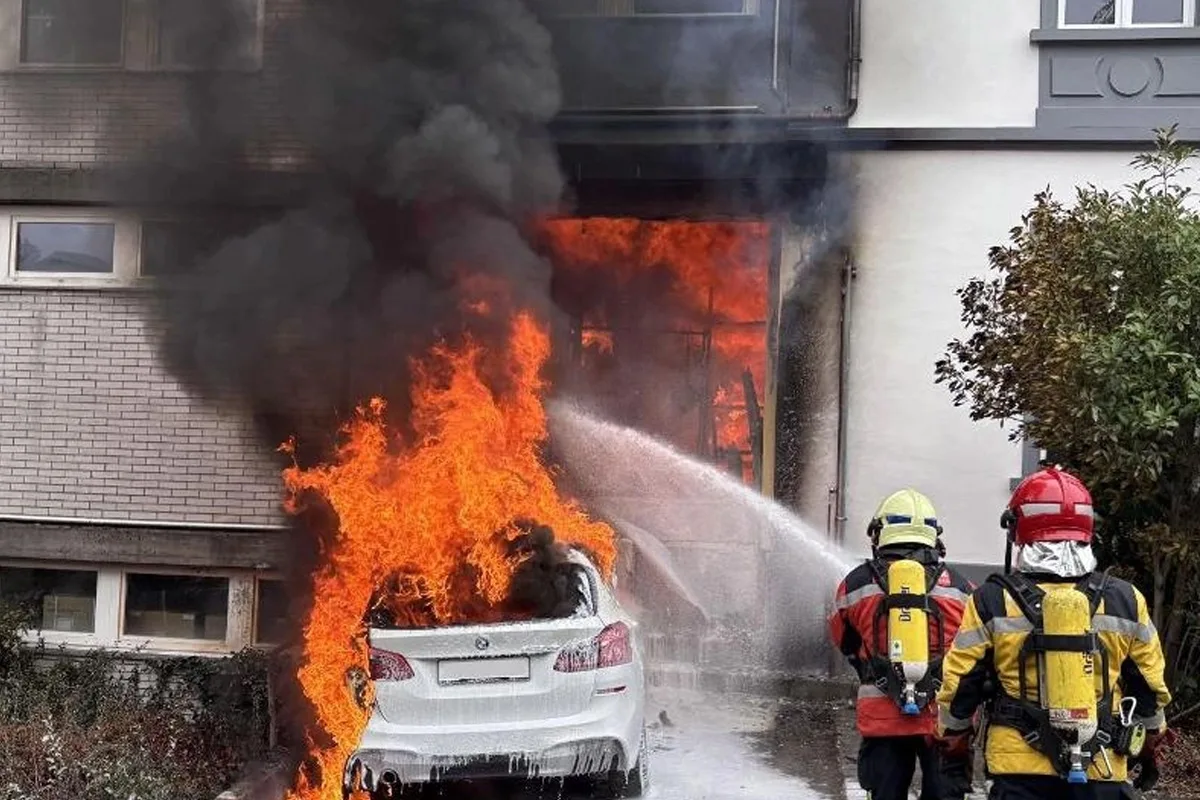 Zwei Feuerwehrleute in Schutzkleidung löschen ein brennendes Auto vor einem Wohnhaus. Dicker Rauch steigt auf.
