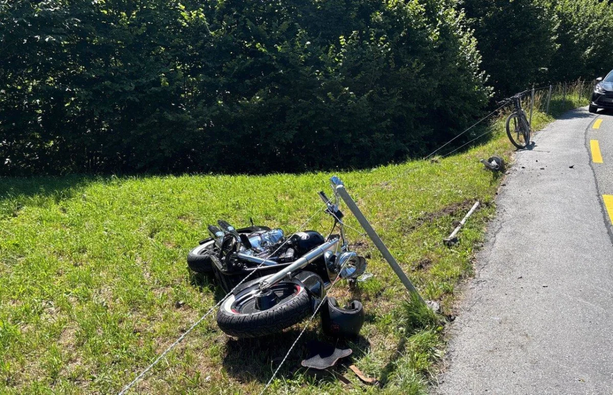 Ein umgestürztes Motorrad liegt im Gras neben einer kurvigen Straße. Ein Fahrrad steht an einem nahegelegenen Drahtzaun.