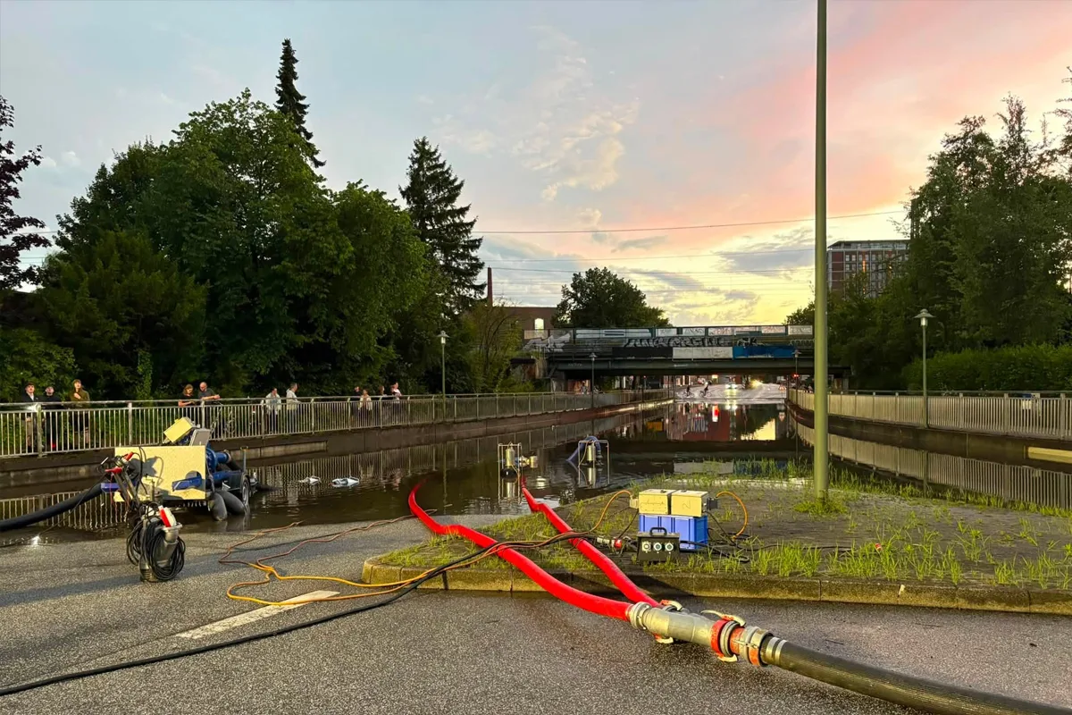 Eine überflutete Straße bei Sonnenuntergang, Schläuche führen zu Pumpen, während Menschen auf einer Brücke stehen.