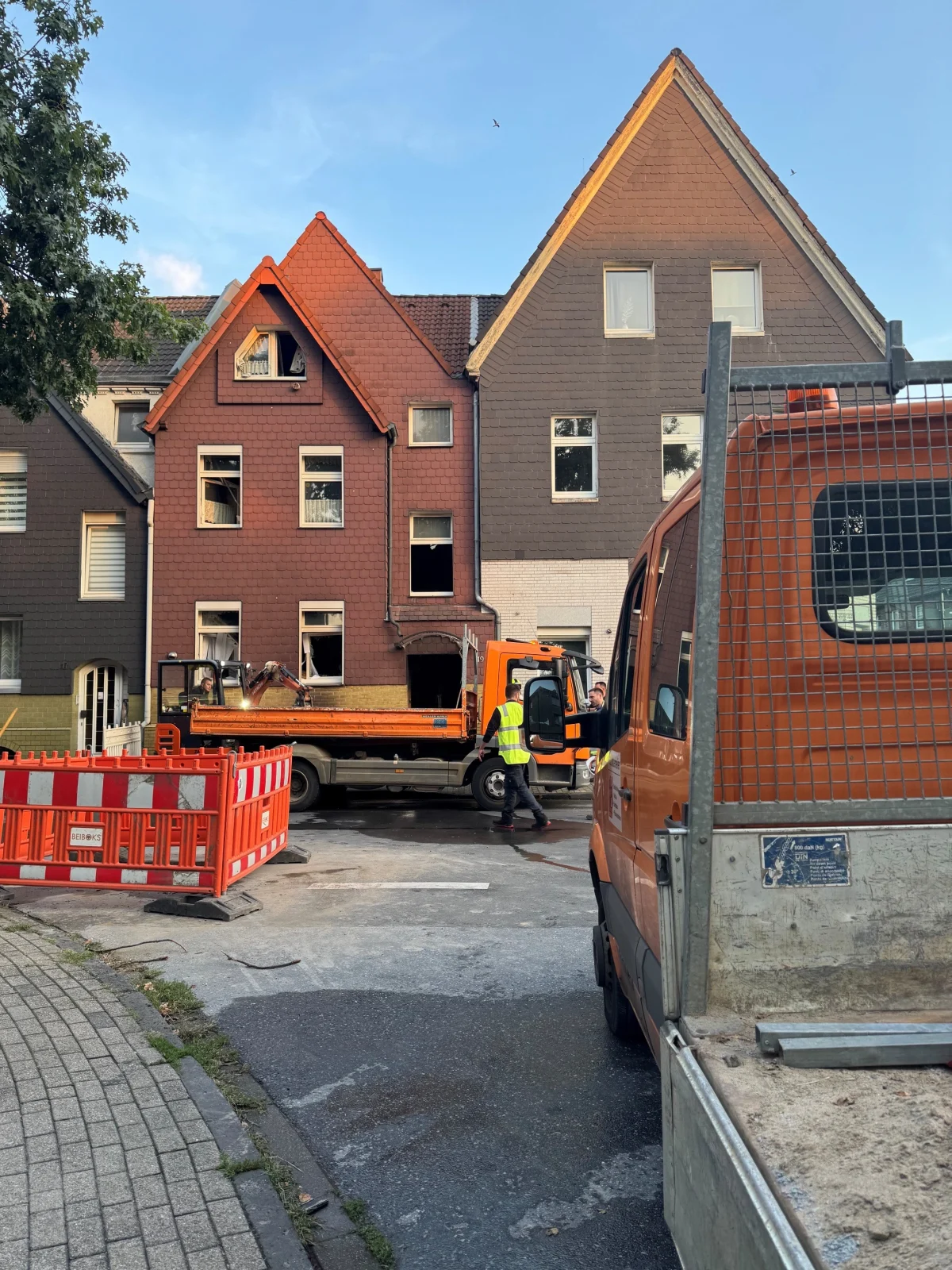 Zwei orangefarbene Lastwagen stehen auf der Straße vor einem Gebäude mit sichtbaren Schäden und offenen Fenstern. Absperrungen sind zu sehen.