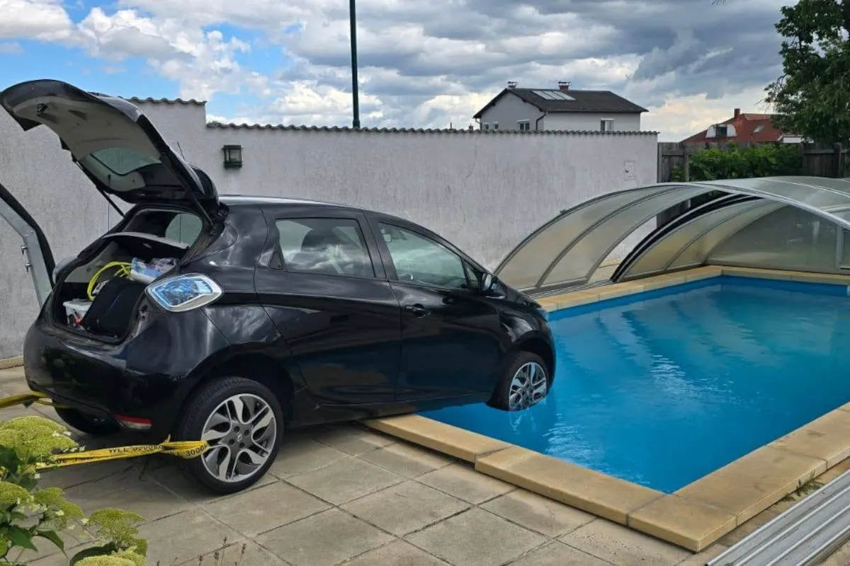 Ein schwarzes Auto ist teilweise in einen Pool gerollt. Der Kofferraum steht offen, und gelbe Kabel sind zu sehen. Der Himmel ist bewölkt.
