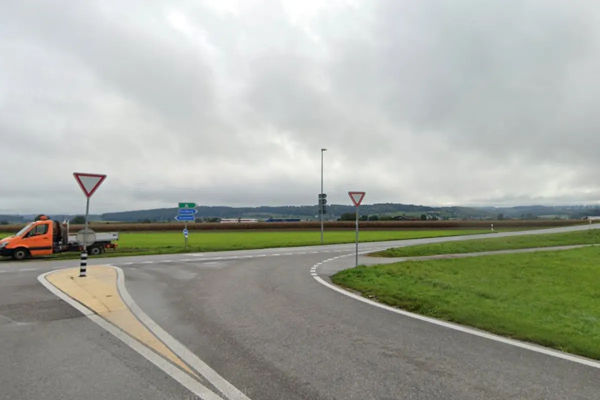 Ein orangefarbener Kleintransporter ist neben einem Verkehrsschild an einem Kreisverkehr abgestellt. Der Himmel ist wolkig und im Hintergrund sind Felder zu sehen.
