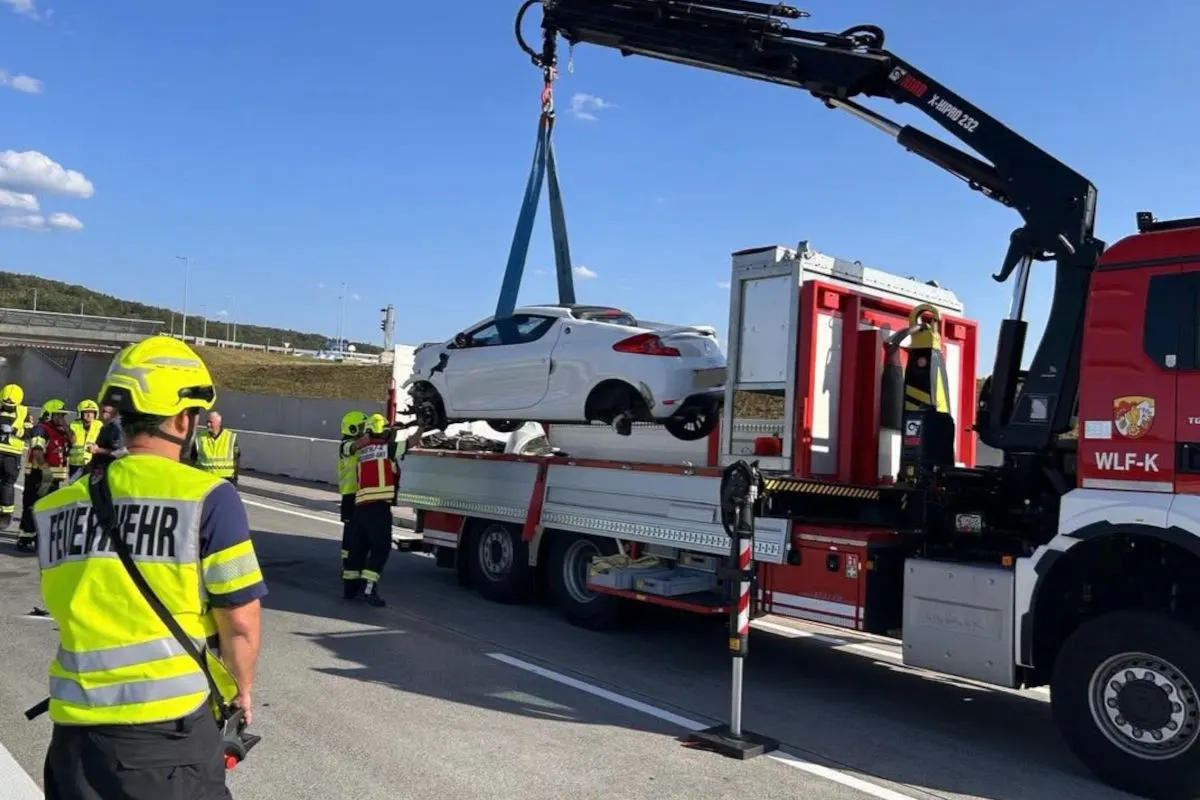 Mehrere Feuerwehrleute bergen ein beschädigtes weißes Auto mithilfe eines Krans auf einer Autobahn bei Tageslicht.