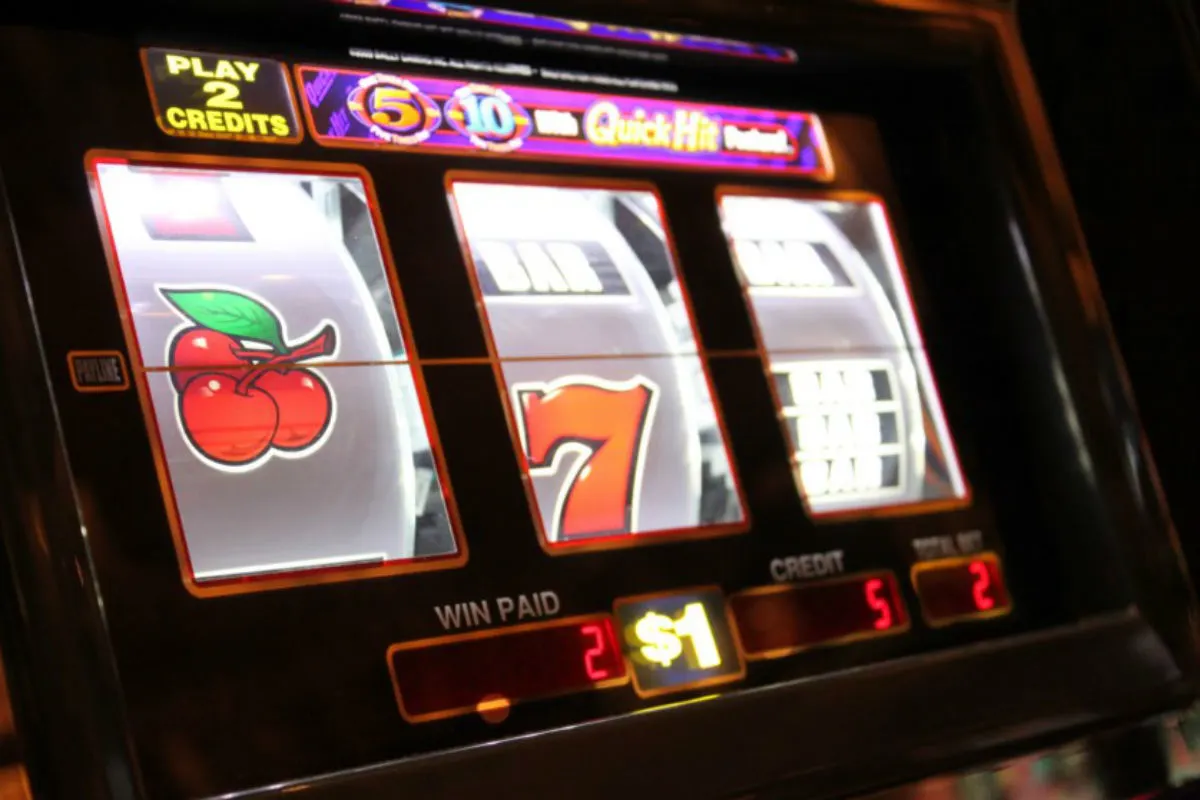 Das Bild zeigt eine Nahaufnahme eines Spielautomaten mit Symbolen wie Kirschen, der Zahl 7 und dem 'Bar'-Symbol. Die Anzeige des Automaten zeigt Gewinne und Einsätze an.