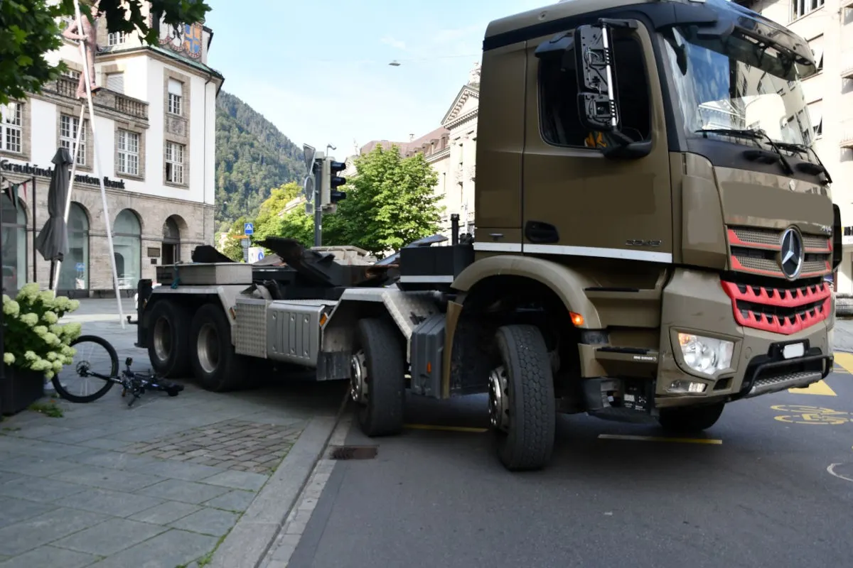 Ein großer Lastwagen steht auf der Straße direkt neben einem umgekippten Fahrrad in einer städtischen Umgebung. Eine Graubündner Kantonalbank ist im Hintergrund zu sehen.