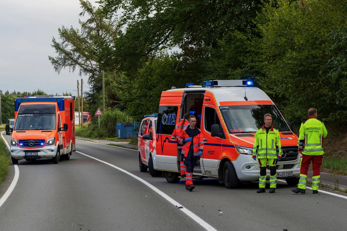 Zwei orange-rote Feuerwehrfahrzeuge stehen auf einer Landstraße, umgeben von Rettungskräften. Dichte Vegetation im Hintergrund.