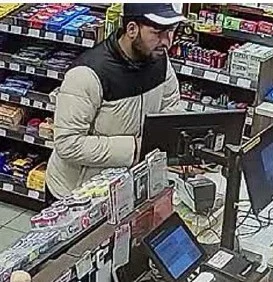 Ein Mann steht an der Kasse eines Geschäfts, umgeben von Regalen mit Waren; die Szene wurde von einer Überwachungskamera aufgenommen.