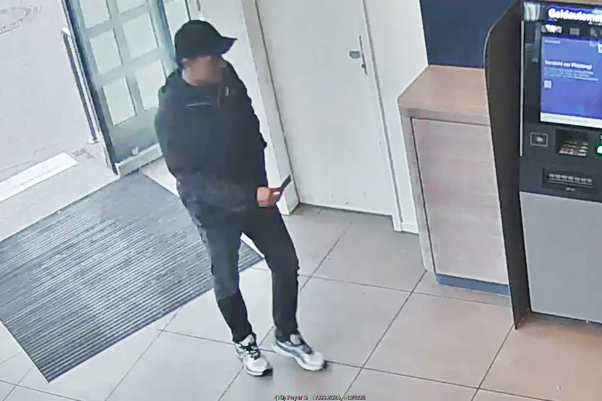 Eine Person mit dunkler Kleidung und Kappe steht in einem Gebäude vor einem Geldautomaten. Der Raum ist einfach und funktional eingerichtet.