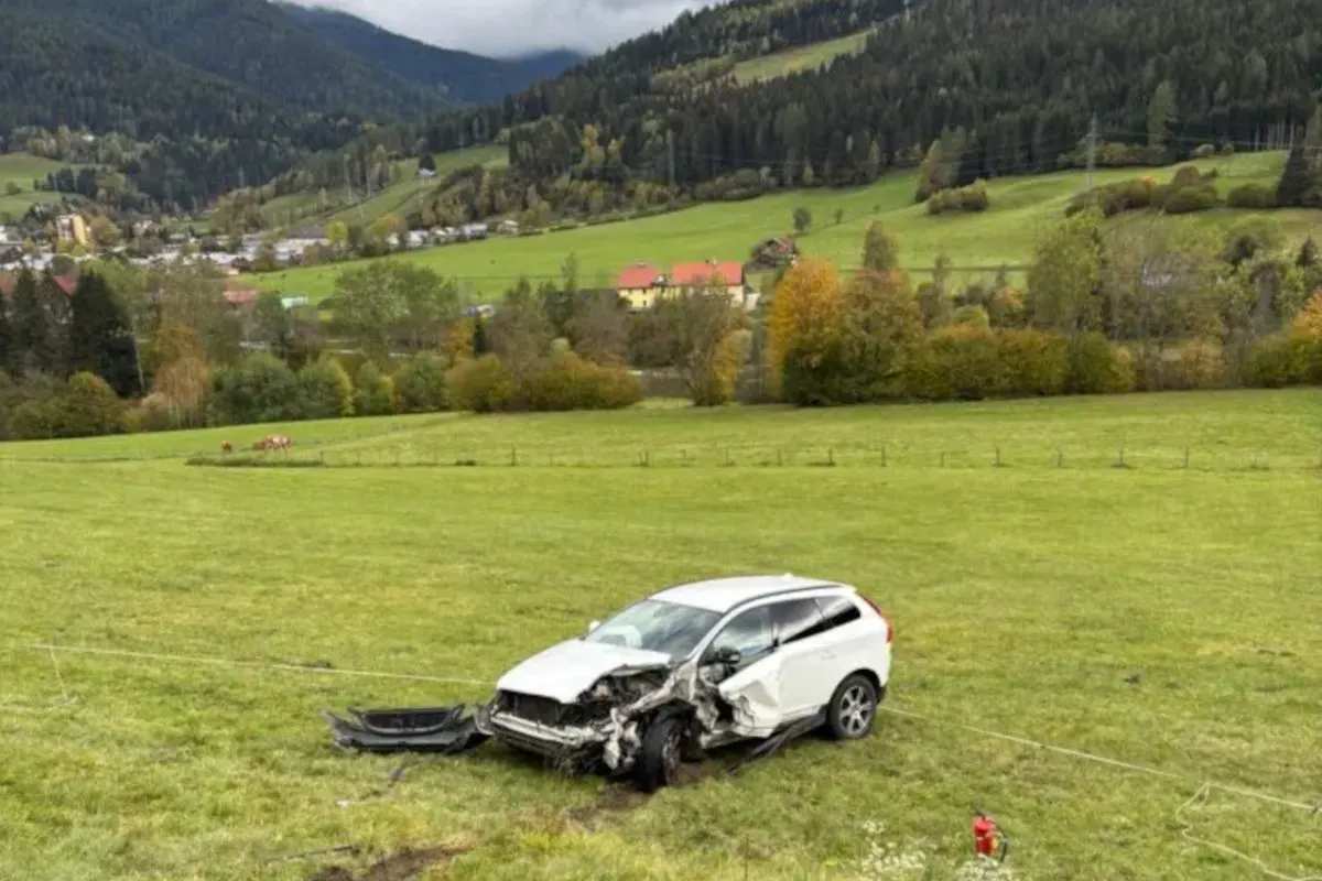 Ein weißes Auto mit schweren Beschädigungen an der Front und der Seite steht auf einer grünen Wiese. Im Hintergrund sind Hügel und eine ländliche Siedlung zu sehen.