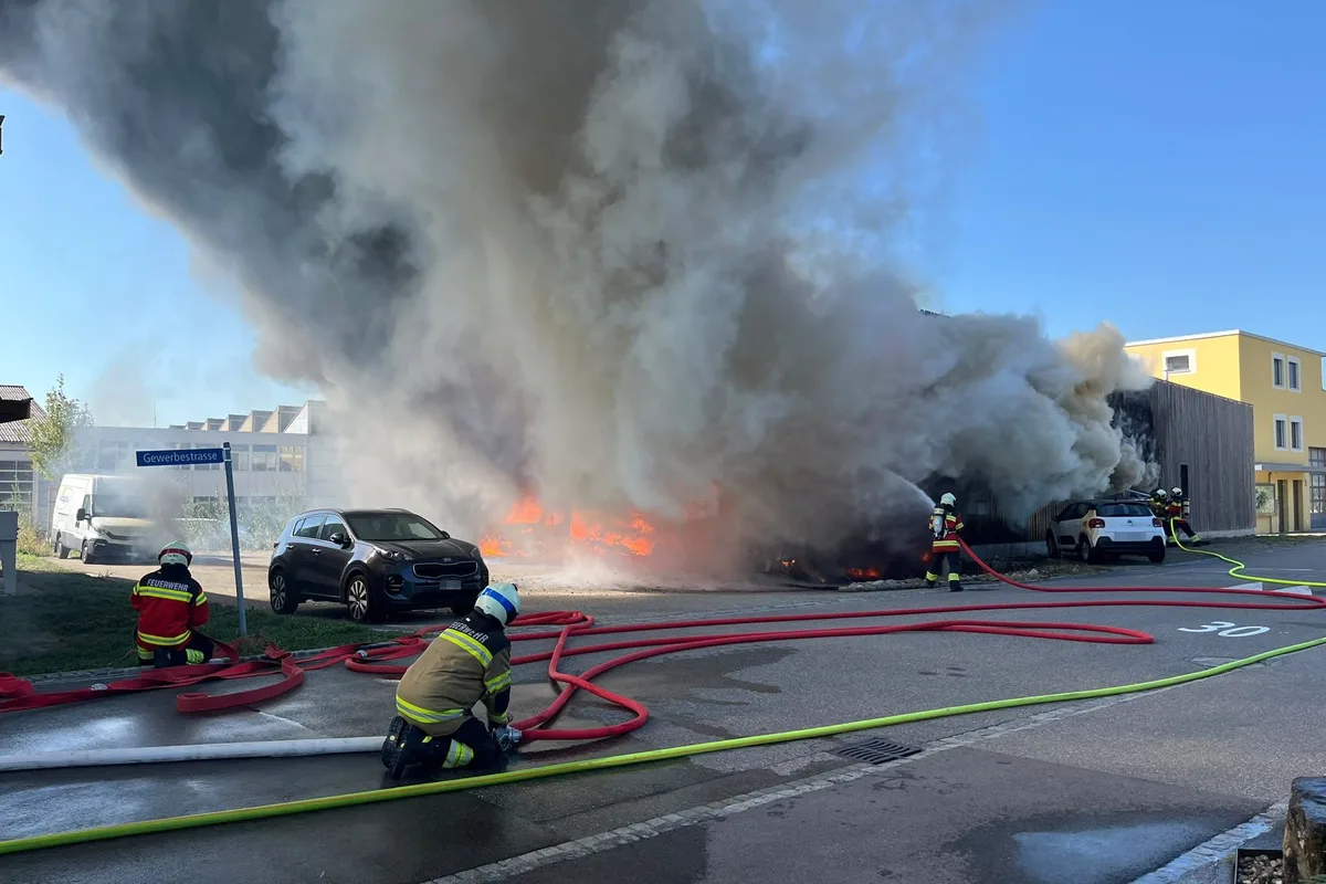 Zwei Feuerwehrleute mit Schutzkleidung sind dabei, mit Schläuchen einen großen Brand an einem Gebäude zu löschen, während dichter Rauch aufsteigt.